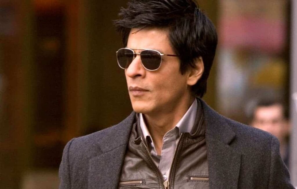 Shah Rukh Khan walking on a busy street, wearing a grey blazer over a light-colored shirt and dark trousers. with blurred figures in the background indicating an urban setting.