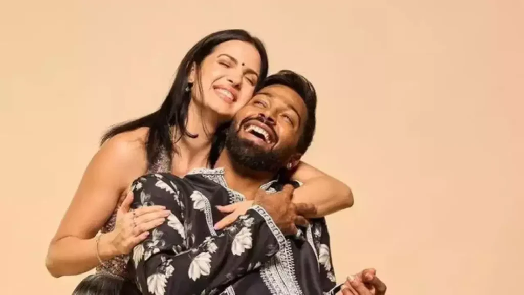 The image features two individuals Hardik Pandya and Natasa Stankovic embracing. One person is seated, while the other stands, leaning over with arms around the seated individual. The standing person wears a black and white patterned dress, while the seated person’s attire isn’t fully visible.