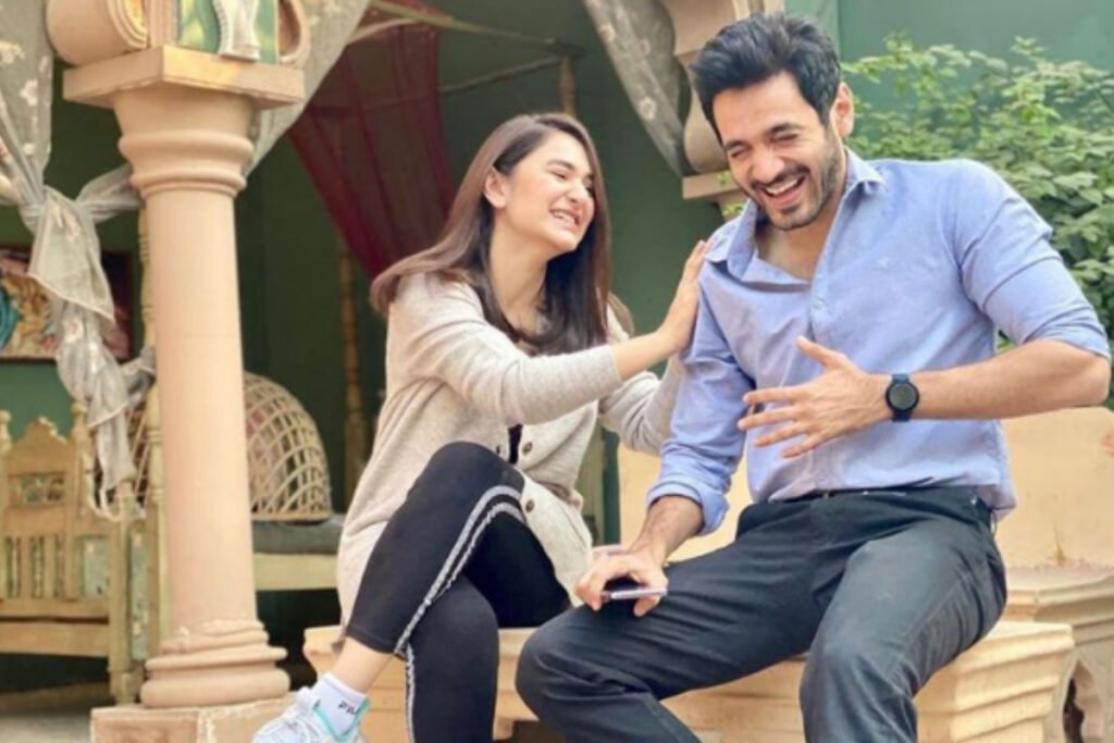 Image shows Yumna Zaidi and Wahaj Ali sitting on a staircase outside a building, engaged in conversation. One is wearing a light grey top and blue jeans, and the other is in a dark shirt and blue jeans.