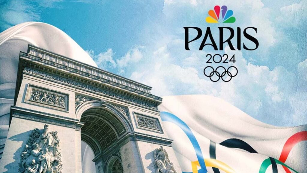 Promotional image featuring the Arc de Triomphe with a large, flowing white fabric draped over it, adorned with colorful Olympic rings. The sky is clear blue with scattered clouds. In the top right corner, there’s the logo for Paris 2024 Olympics, which includes the Olympic rings and the NBC peacock logo.