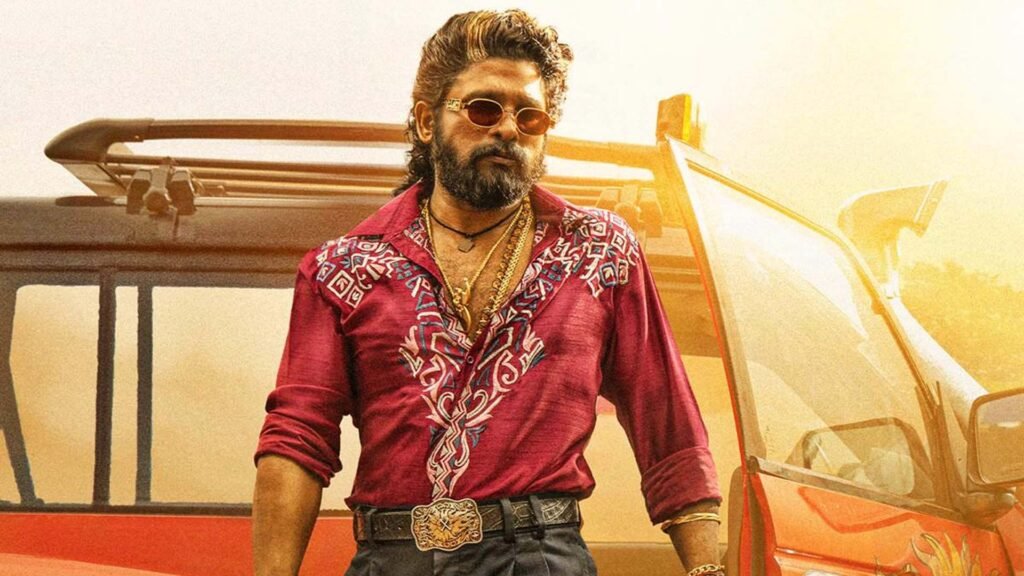 Allu Arjun standing confidently in front of a red tractor, dressed in a maroon patterned shirt and dark trousers with an elaborate belt buckle, set against a rural backdrop.