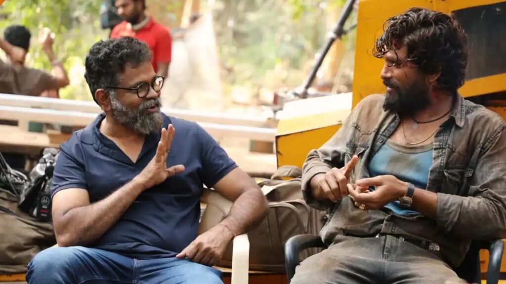 The image features two individuals seated next to each other. Promotional photo capturing a behind-the-scenes moment between Allu Arjun and Sukumar. Allu Arjun, known as the ‘Stylish Star,’ wears a dark blue shirt, while Sukumar, the acclaimed director, is dressed in layered attire. The setting suggests an outdoor film set, with equipment and greenery in the background.