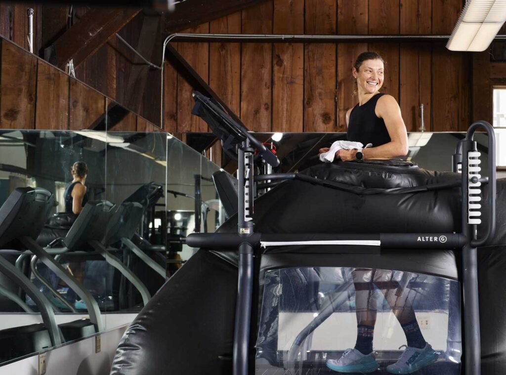 Individual using an AlterG Anti-Gravity Treadmill at a gym. The treadmill is designed to reduce weight-bearing stress during physical therapy or training, featuring a unique air pressure chamber that supports the user comfortably around the lower body. The individual is focused on their workout, wearing athletic attire and running shoes, with reflections visible in the surrounding mirrors.