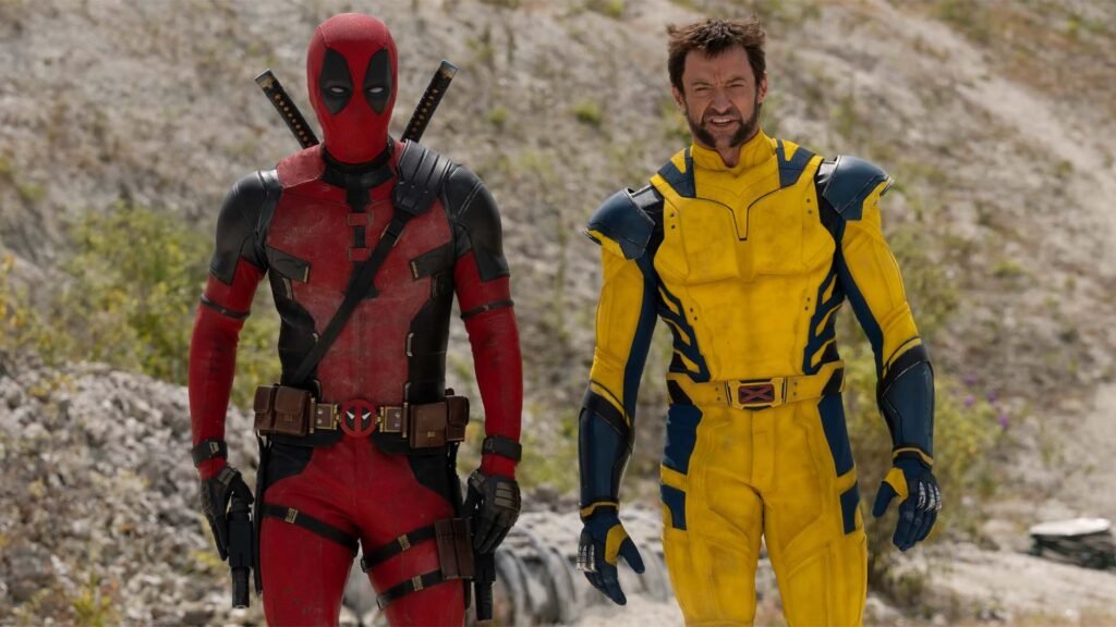 The image features two characters standing side by side. On the left is a character in a red and black suit with a mask, representing Deadpool. On the right is another character in a yellow and blue suit with their face obscured, intended to represent Wolverine. They appear to be standing on rocky terrain with sparse vegetation in the background. This image may be relevant for fans of comic book characters or movies, showcasing two popular Marvel characters known for their distinctive costumes and association with the X-Men series.