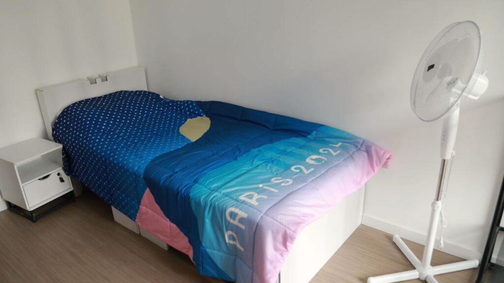 Image of a neatly arranged bedroom featuring a single bed with a blue and pink duvet cover that has ‘Paris 2021’ printed on it. A white bedside table, a white headboard, and an electric fan are also visible, contributing to a clean and simple room decor.