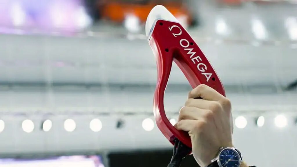 Close-up of a person’s hand holding a red Omega starting block against an indoor blurred background, likely at a sporting event where precision timing is essential.