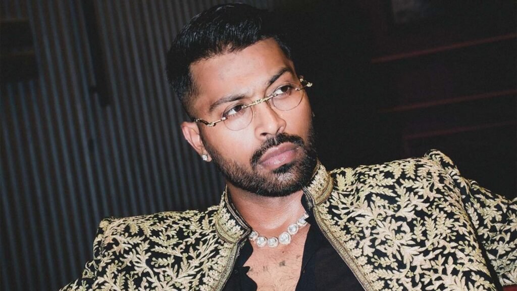 The image depicts an individual wearing a black shirt with a deep neckline and an ornate, gold-patterned jacket. The person is also adorned with what appears to be a pearl necklace. The fashion statement suggests a bold and luxurious style, which could be associated with Hardik Pandya’s flamboyant persona off the cricket field.