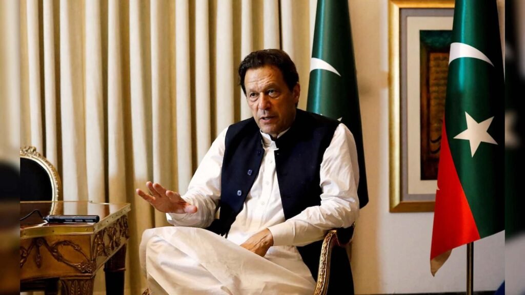 Imran Khan, wearing a dark waistcoat over a light-colored traditional shalwar kameez, seated on a chair with a blurred face, gesturing with his right hand. The background features cream-colored curtains and the national flag of Pakistan.