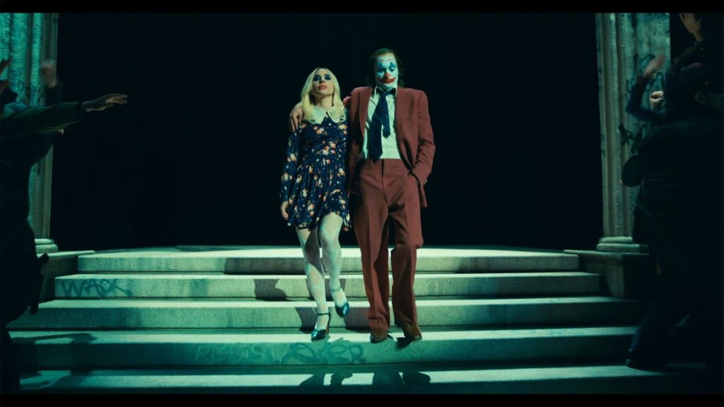 The image is a still from the official trailer for “Joker: Folie À Deux.” It features two characters, one wearing a floral dress and the other in a red suit, both with their faces obscured by white blocks to maintain anonymity. They are standing on steps with their arms interlocked, suggesting a dance or performance. The setting appears to be theatrical with dramatic lighting and figures around them that seem to be part of an audience or ensemble cast. This image is relevant as it captures a moment from the trailer that may be significant for its plot or character development.