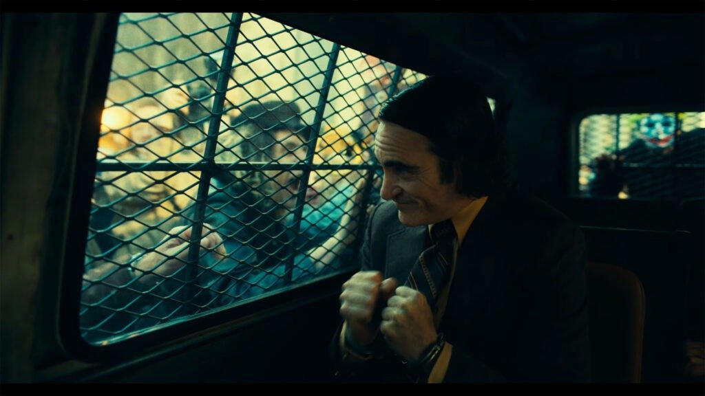 The image is a still from the official trailer for “Joker: Folie À Deux.” It features an individual presumed to be Arthur Fleck, who is inside a vehicle looking out through a window with metal gridwork. The character’s face is obscured by an anonymizing gray box, likely added for privacy or copyright reasons. The lighting is dim, and the colors are muted, suggesting a somber or intense mood typical of the Joker narrative. This image captures a moment from the trailer that may be significant for its plot or character development.