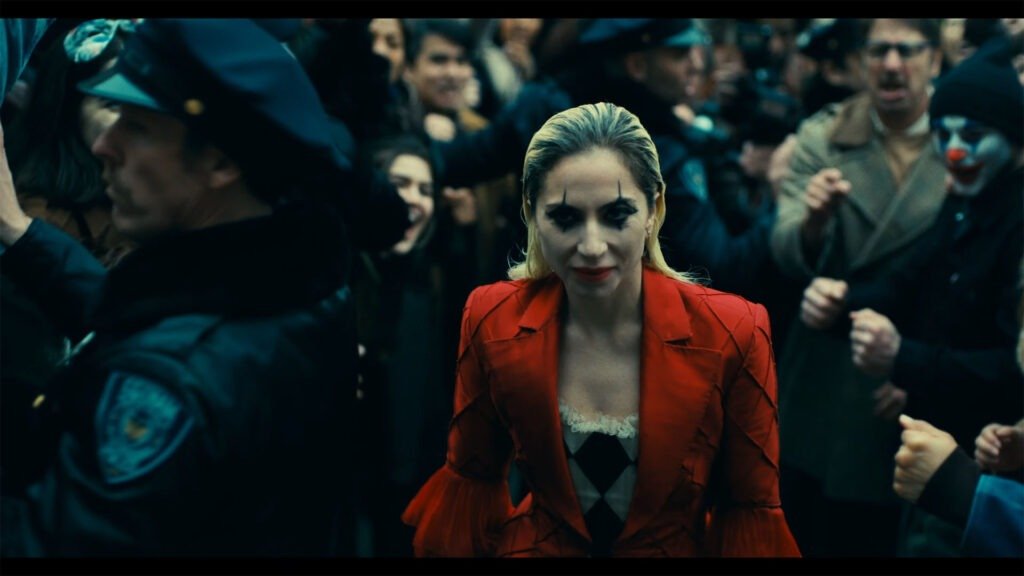 A central figure with blonde hair is dressed in a red jacket and a sparkling silver top at an outdoor event. The individual stands out amidst a crowd of people whose faces have been obscured by gray blocks. The setting suggests an exciting or chaotic gathering, possibly during a pivotal moment in the film. Lady Gaga portrays Harley Quinn in this anticipated sequel, which explores the twisted relationship between Joker (played by Joaquin Phoenix) and Harley Quinn.