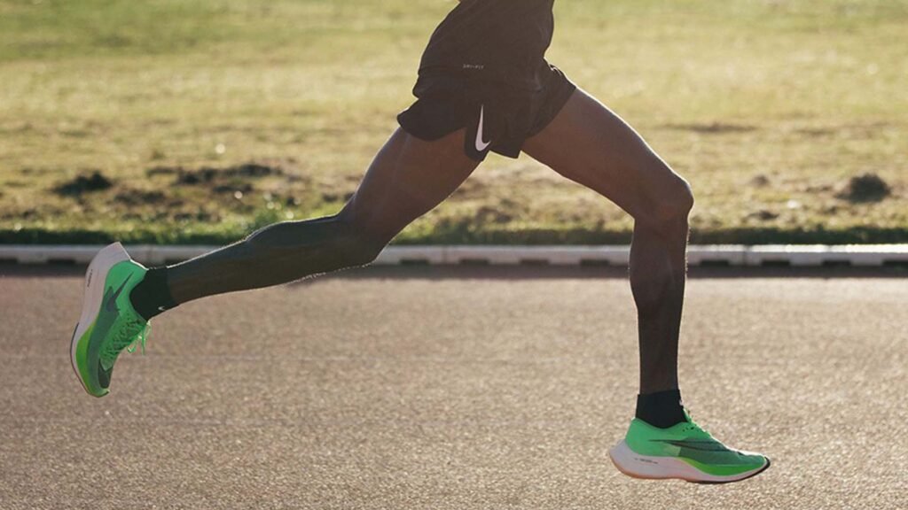 The image shows marathon champion Eliud Kipchoge in mid-stride during a race. The focus is on his lower body, highlighting his toned leg muscles as he runs. He is wearing black shorts and bright green running shoes, which stand out against the blurred background of a road and grassy area. This image captures the dynamic motion and physicality of marathon running, showcasing Kipchoge’s athletic prowess and endurance.