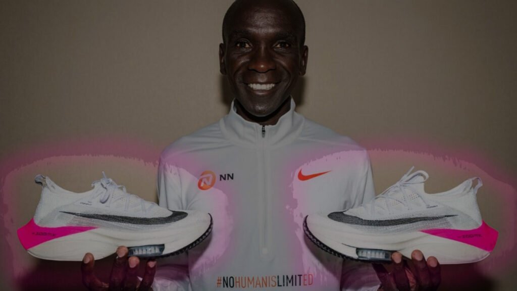 The image shows marathon champion Eliud Kipchoge holding a pair of Nike running shoes, one in each hand. The shoes are predominantly white with pink and black accents. Kipchoge is wearing a white jacket with the NN logo on the left side and a Nike swoosh on the right, symbolizing sponsorship. Across the chest of the jacket, the hashtag #NOHUMANISLIMITED is prominently displayed.