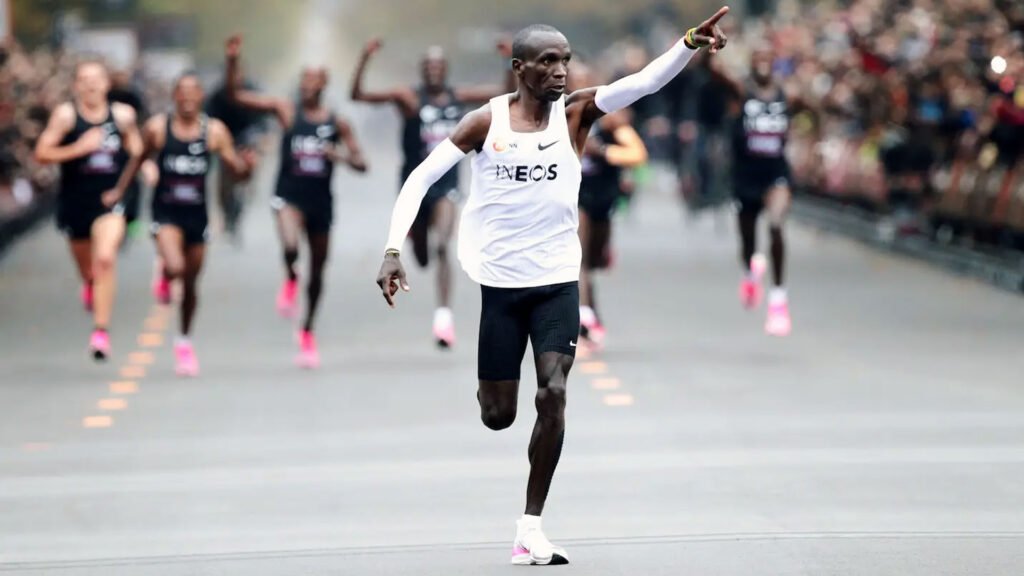 The image shows marathon champion Eliud Kipchoge in mid-stride during a race. He is wearing a white singlet with the logo “INEOS” and black shorts. His right arm is extended forward, pointing ahead with one finger, suggesting determination or signaling a victory. The background features blurred spectators on either side of the barricaded course, cheering on the runners. Several other runners, wearing pink shoes, are visible behind Kipchoge, following him in the race.