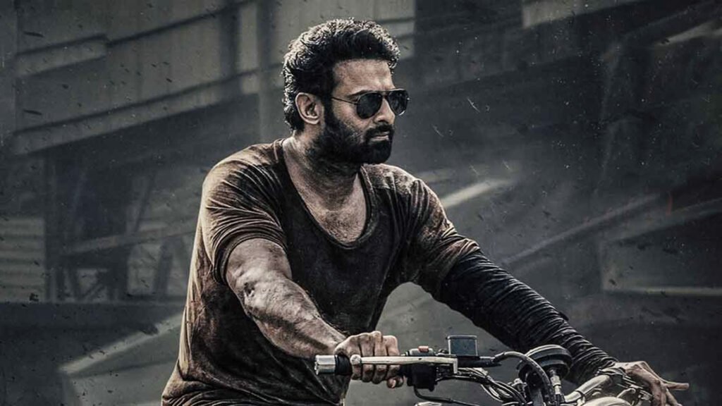 The image shows an individual riding a motorcycle. The person wears a dirtied, dark-colored t-shirt and appears to be in motion, as suggested by the dynamic pose and blurred background, indicating speed. The setting resembles an industrial or possibly post-apocalyptic environment, with grey tones and debris visible. This image could be relevant for representing the character played by Prabhas in a specific context or movie scene Salaar.