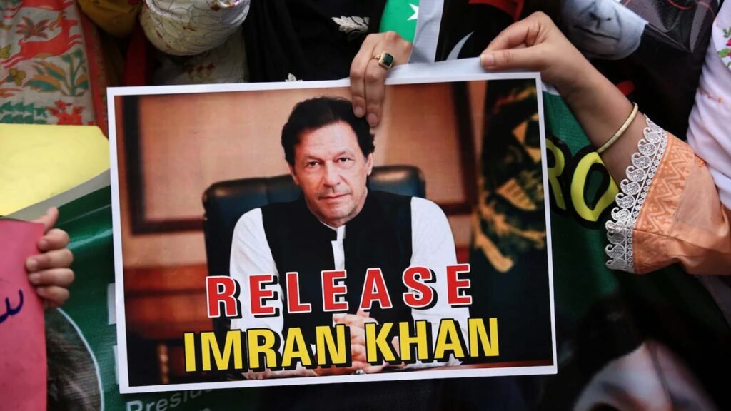 Poster with the text ‘RELEASE IMRAN KHAN’ in bold white and red letters, featuring an individual, presumably Imran Khan, sitting in an official setting with a blurred face for privacy reasons.