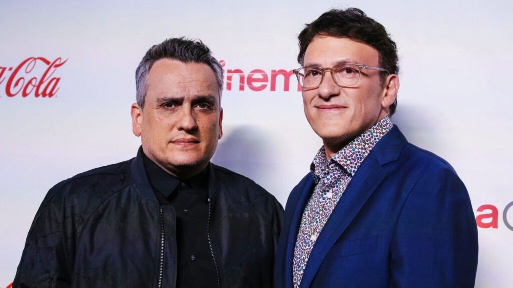 The image features two individuals, identified as the Russo Brothers, standing side by side. The individual on the left is wearing a black jacket over a black shirt, and the one on the right is dressed in a patterned blue jacket over a white shirt with an open collar. Both are facing forward and appear to be at an event with a red background that includes logos, one of which reads “Coca-Cola.”