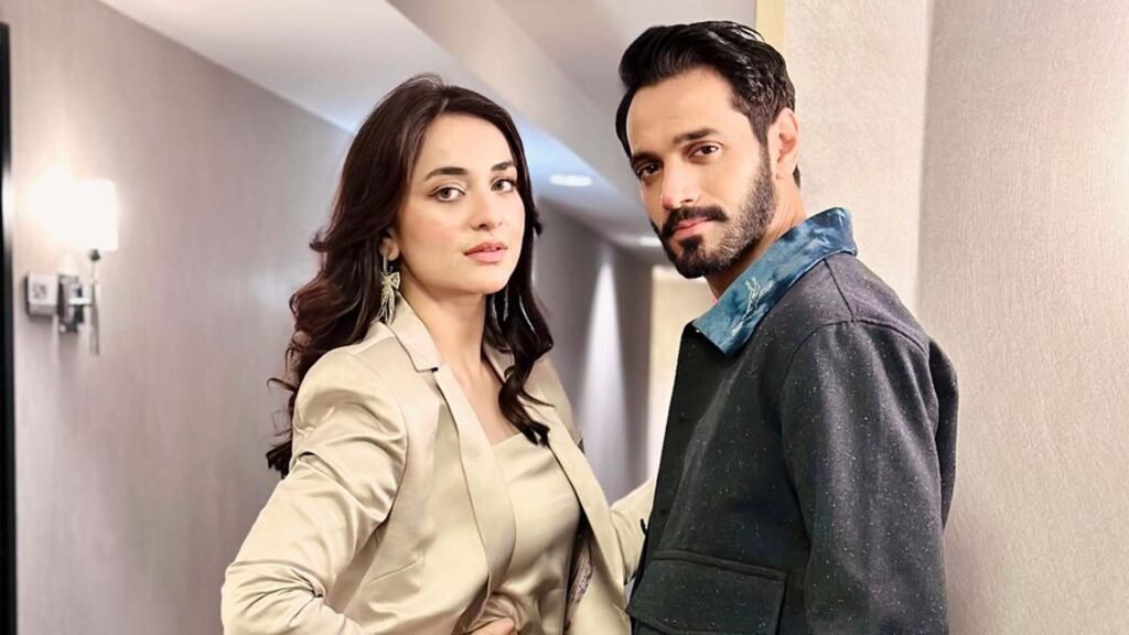 Image shows Yumna Zaidi and Wahaj Ali standing side by side against a neutral-toned wall. Yumna Zaidi is wearing a light beige blazer over a white top, and Wahaj Ali is in a dark jacket with a textured collar.