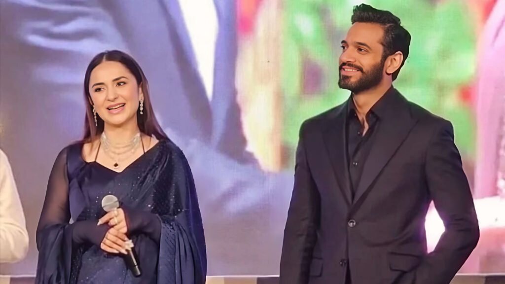 Yumna Zaidi and Wahaj Ali at an event. Yumna Zaidi is seated, wearing a dark blue outfit with embellishments, holding a microphone. Wahaj Ali stands beside her, dressed in a black suit. Both are facing towards an audience, with a colorful blurred background suggesting the setting of a stage or festive occasion.