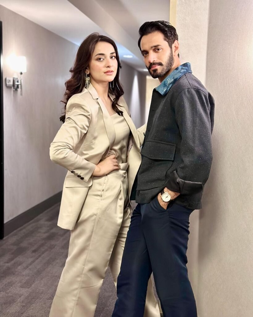 Yumna Zaidi and Wahaj Ali, standing side by side in a hallway with confident postures.