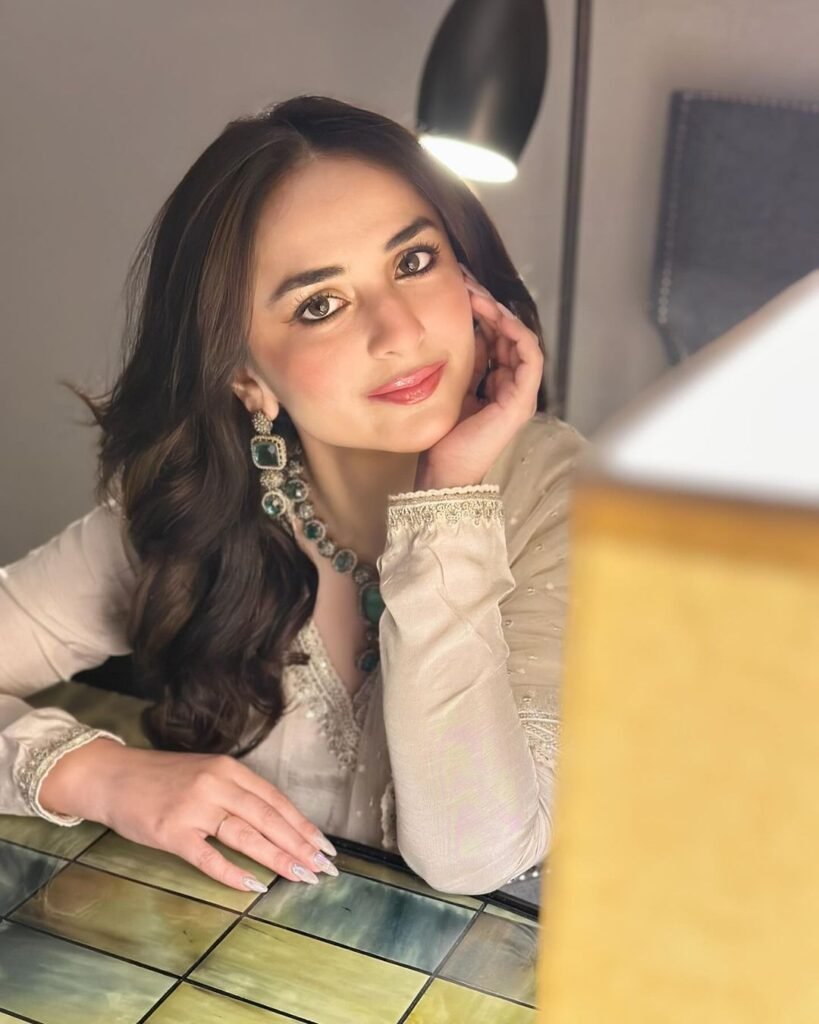 Yumna Zaidi seated at a table, wearing a light-colored outfit with embroidery and a green necklace. Her face is partially obscured by her hand in a thoughtful pose. The table has a reflective surface with multicolored square patterns, creating a casual and relaxed atmosphere.