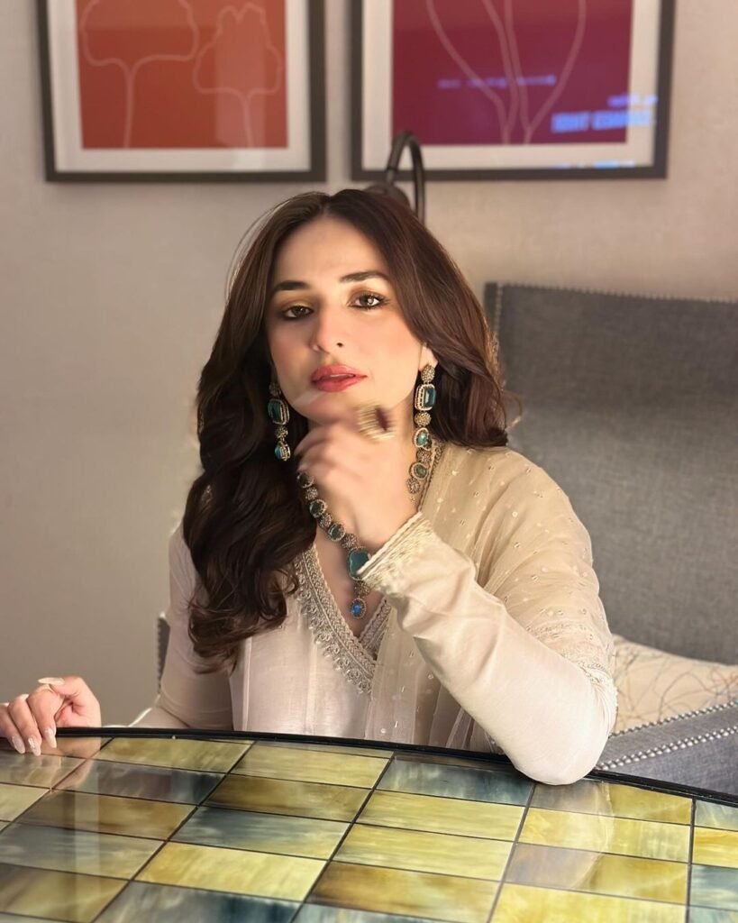 Yumna Zaidi seated at a table, wearing a white outfit with intricate embroidery. She is accessorized with large, dangling earrings and a matching necklace with green accents. The background features a cushioned bench and abstract art pieces in warm tones.