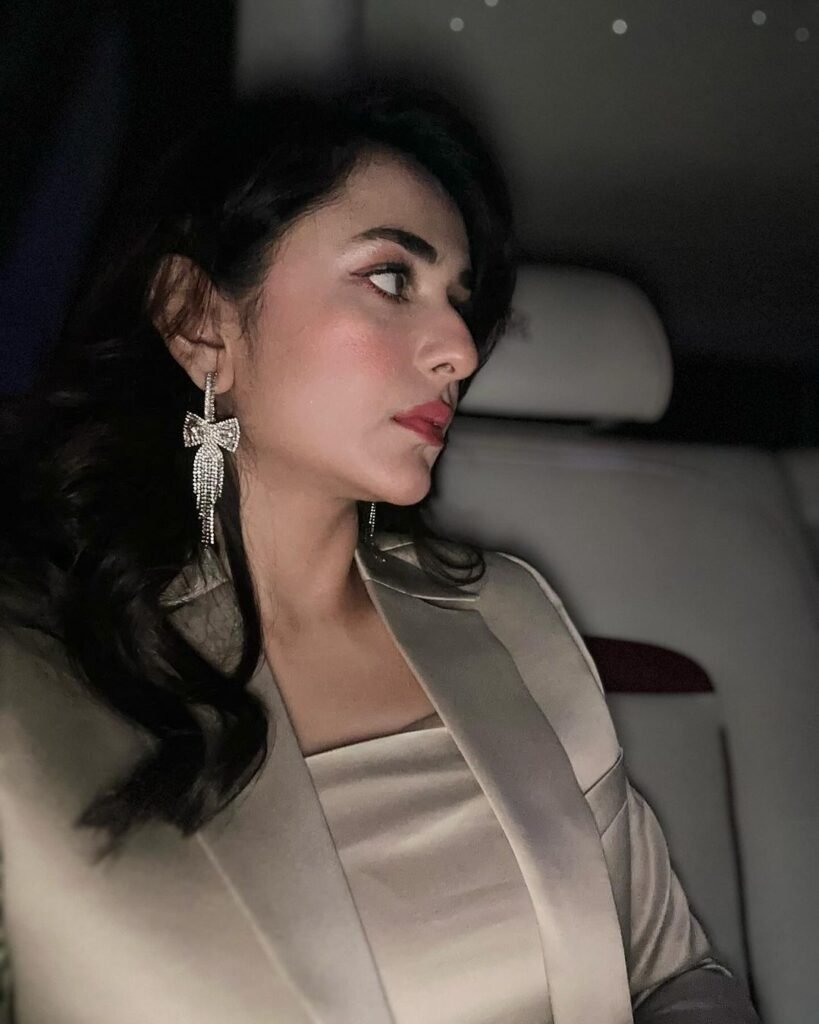 Yumna Zaidi, a renowned Pakistani actress, seated in a vehicle. She is wearing a light-colored outfit with a lapel and has dark, wavy hair adorned with a dangling earring. The interior of the vehicle is dimly lit, with small lights visible in the background, possibly part of the car’s interior or external lights.