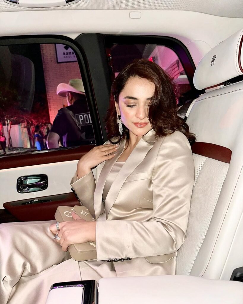 Yumna Zaidi, a well-known Pakistani actress, seated in the back of a car with a cream interior. She is dressed in a light-colored suit and holding a small clutch or purse. Outside the car window, there are people gathered, including a figure in police attire, suggesting that she might be attending a public event or gathering.