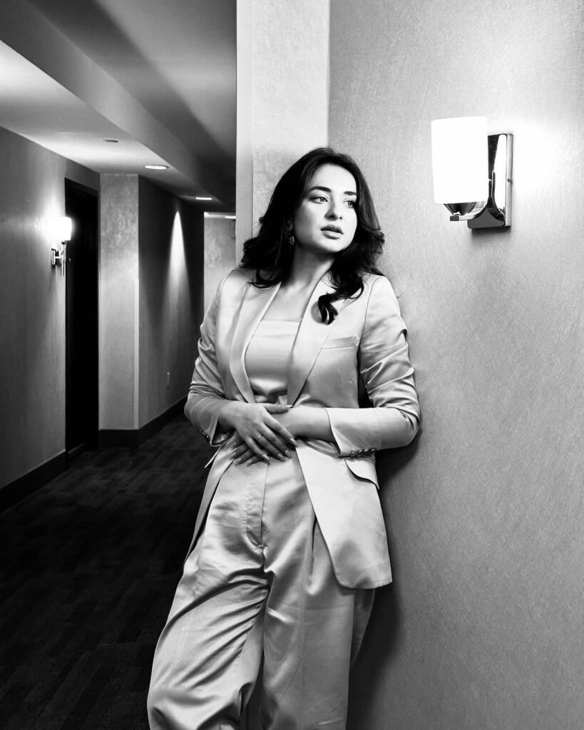 Yumna Zaidi, a renowned Pakistani actress, standing in a corridor. She is dressed in a stylish, light-colored long coat paired with matching trousers. The corridor is well-lit with elegant wall-mounted lights, creating a sophisticated ambiance.