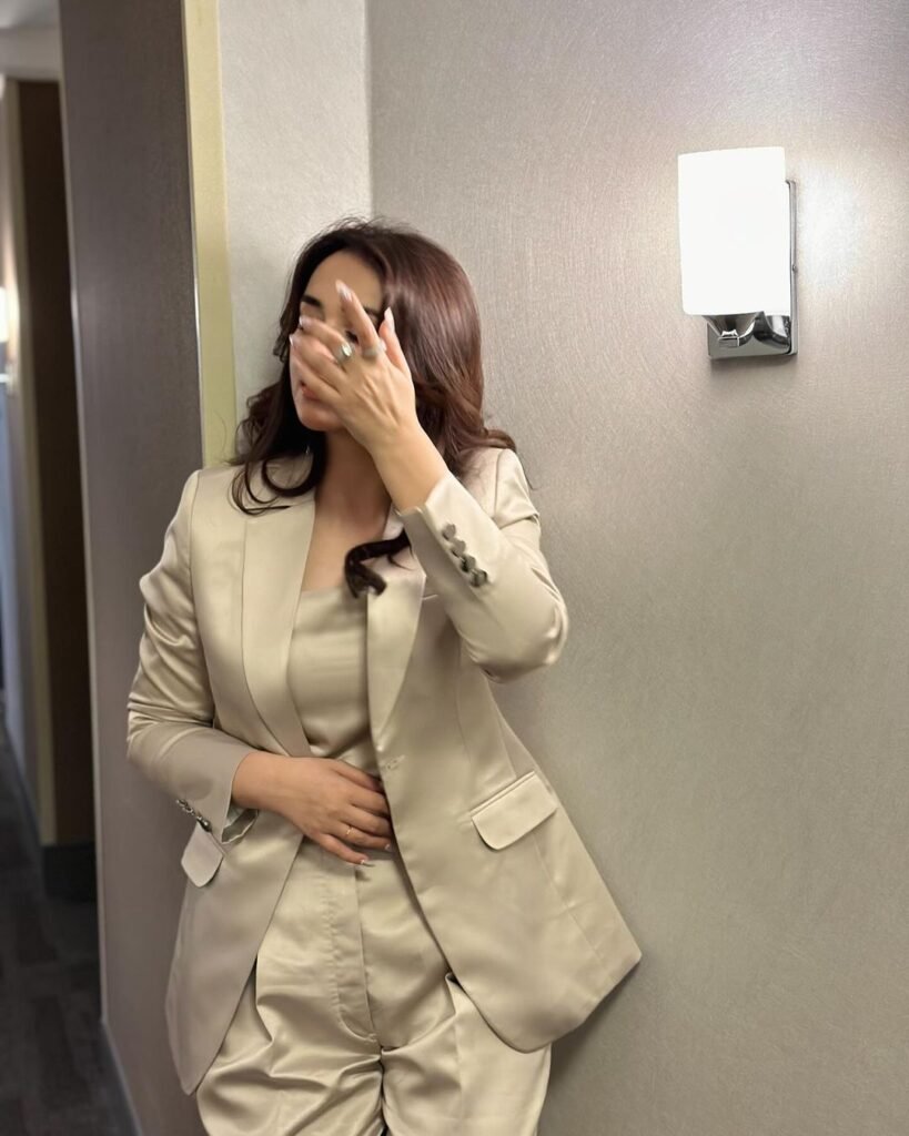 Yumna Zaidi standing in a corridor next to a door, wearing a light-colored suit and holding something close to her chest with both hands. There is a wall-mounted light fixture above her on the right side.