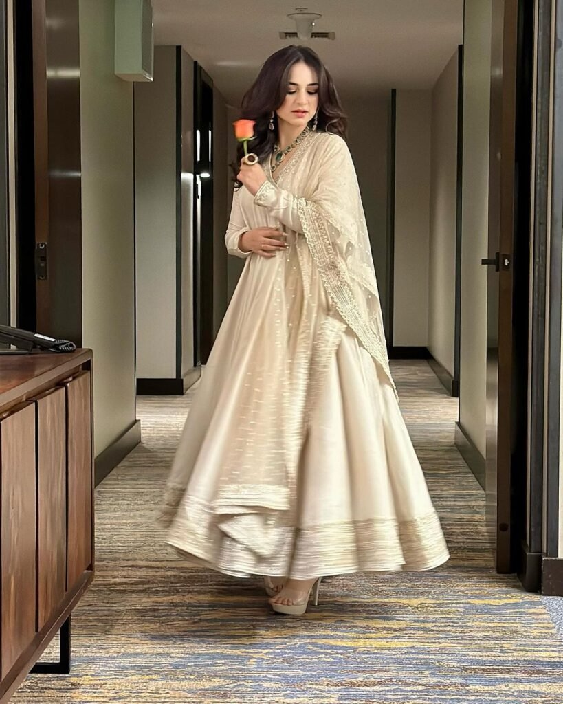 Yumna Zaidi standing in a hotel corridor, wearing a full-length beige dress with intricate embroidery. She is holding a smartphone and appears to be taking a selfie. The corridor has patterned carpeting, wood-paneled walls, and modern lighting fixtures.