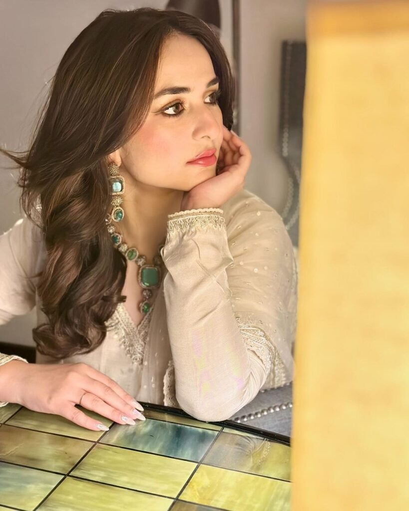 Yumna Zaidi seated at a table, wearing a white outfit with intricate embroidery and a green necklace. Her long, wavy hair cascades down her shoulders. The setting appears to be indoors, with reflective surfaces and warm lighting creating a cozy atmosphere.