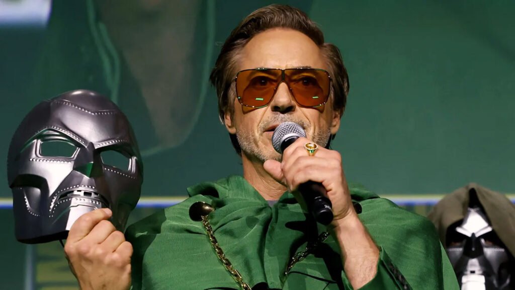 Image of an unidentified person at an event, speaking into a microphone while wearing sunglasses and a green hooded outfit, with their face pixelated for privacy. A blurred square obscures part of the image on the left, and a metallic arm or gauntlet is partially visible on the right.