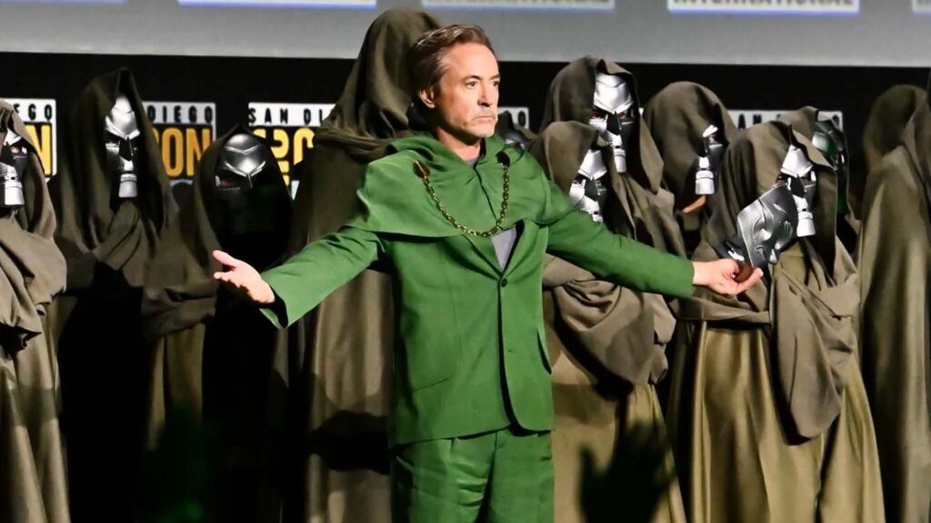 The image shows an individual standing on a stage with arms outstretched, wearing a green suit and metallic gloves. The face is obscured by a brown rectangle, making identification impossible. Behind the figure are several individuals in cloaks with metallic masks, resembling Dr. Doom’s iconic look from the comics. This image could be relevant for illustrating a costume event or a comic book convention where attendees dress as characters from comic books, such as Dr. Doom.