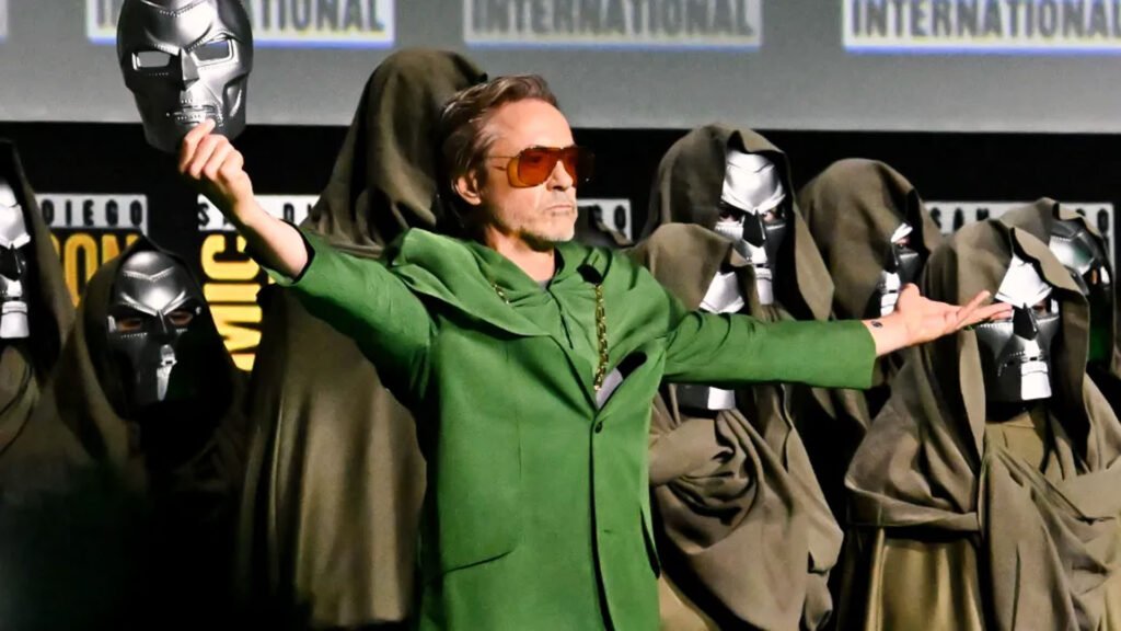Robert Downey Jr., a famous actor known for his role as Iron Man. He is standing on stage at a Comic-Con International event, extending his arms wide with a smile. Behind him, several individuals are dressed in costumes resembling characters from the ‘Iron Man’ movies, including cloaks and masks of Iron Man’s helmet.