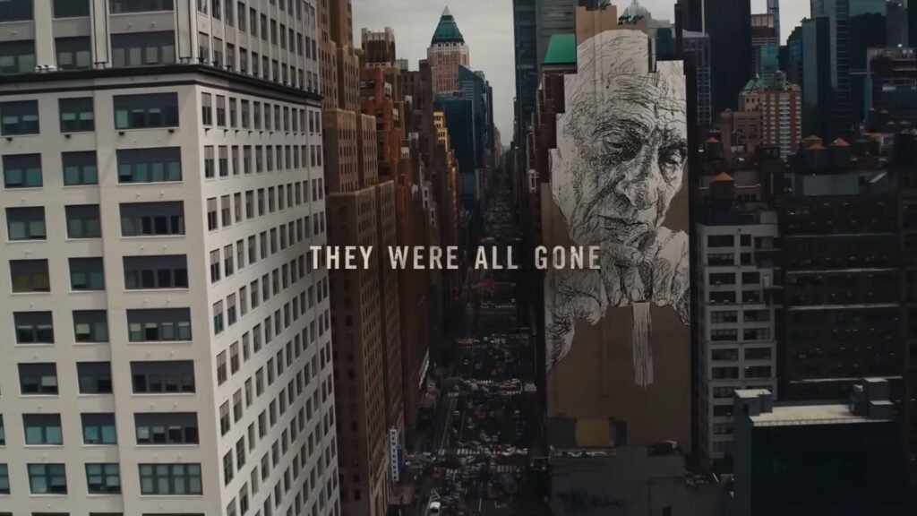 The image depicts an aerial view of a cityscape with high-rise buildings lining the street. One building features a large mural of an elderly person’s face. Overlaid on the image is the text “THEY WERE ALL GONE.”