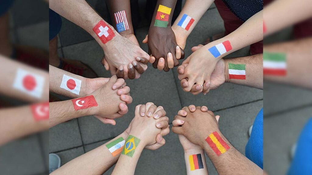 The image shows a group of hands joined together in the center of the frame, forming a circle. Each hand has a different flag painted on its wrist or back, representing various countries such as Switzerland, Japan, Ghana, Italy, Turkey, Brazil, Germany, and Belgium. This image visually represents unity and diversity through the symbolic gesture of joined hands from individuals presumably from different nations.