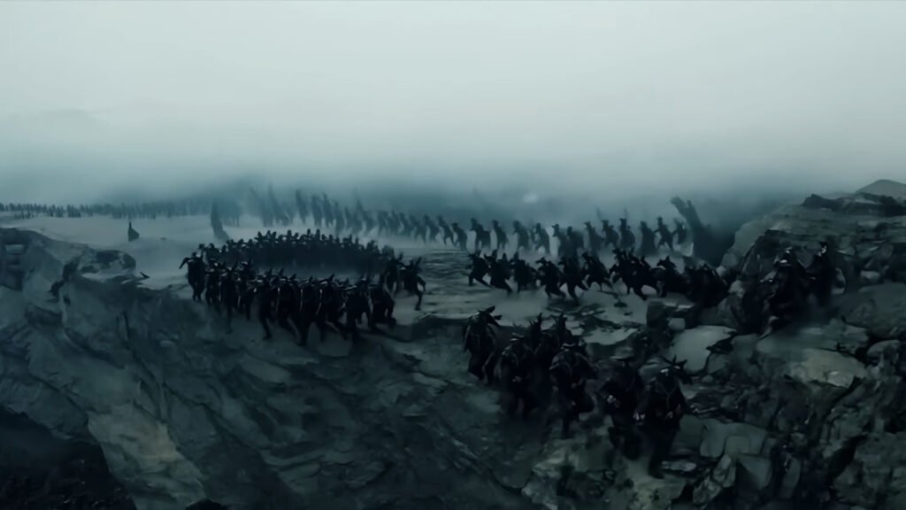 a large group of armored soldiers standing on the edge of a vast canyon. The atmosphere is bleak and foggy, creating an ominous mood. The soldiers are arranged in formation, suggesting preparation for an epic battle or significant event. This scene is from the movie “Gog and Magog,” capturing the anticipation and scale of the narrative.

