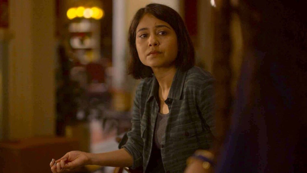 Person identified as Golu from ‘Mirzapur’ seated at a table during an indoor scene with warm ambient lighting, engaged in conversation in Mirzapur Bonus Episode.