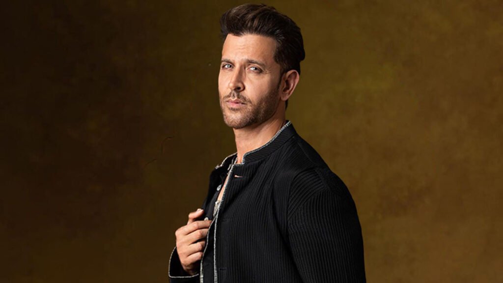 Hrithik Roshan standing in profile view against a brown textured background. He is wearing a dark-colored, textured blazer with visible buttons and appears to be holding onto the lapel of the blazer. The face is not visible as it is obscured by pixelation for privacy. This image conveys a sense of sophistication and style, suitable for a website featuring Hrithik Roshan.