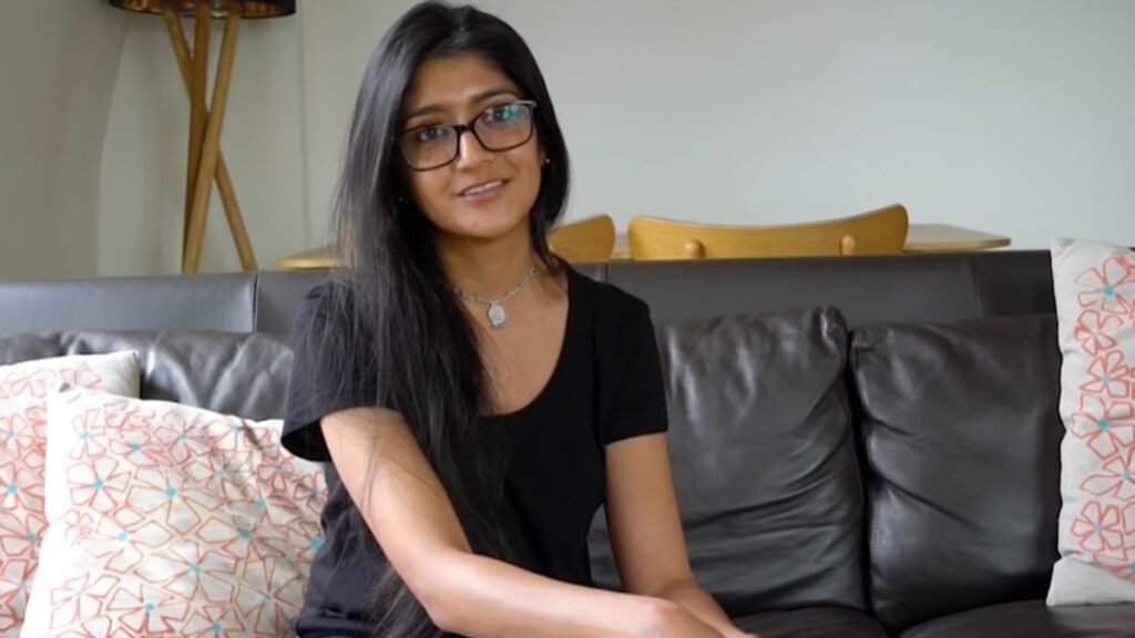 The image shows Laraib Atta, a Pakistani visual effects (VFX) artist, seated on a dark-colored leather sofa with various patterned cushions. She is wearing a black top and has dark hair. Behind her, there is a wooden chair with a light-colored seat and a floor lamp with a slender stand.