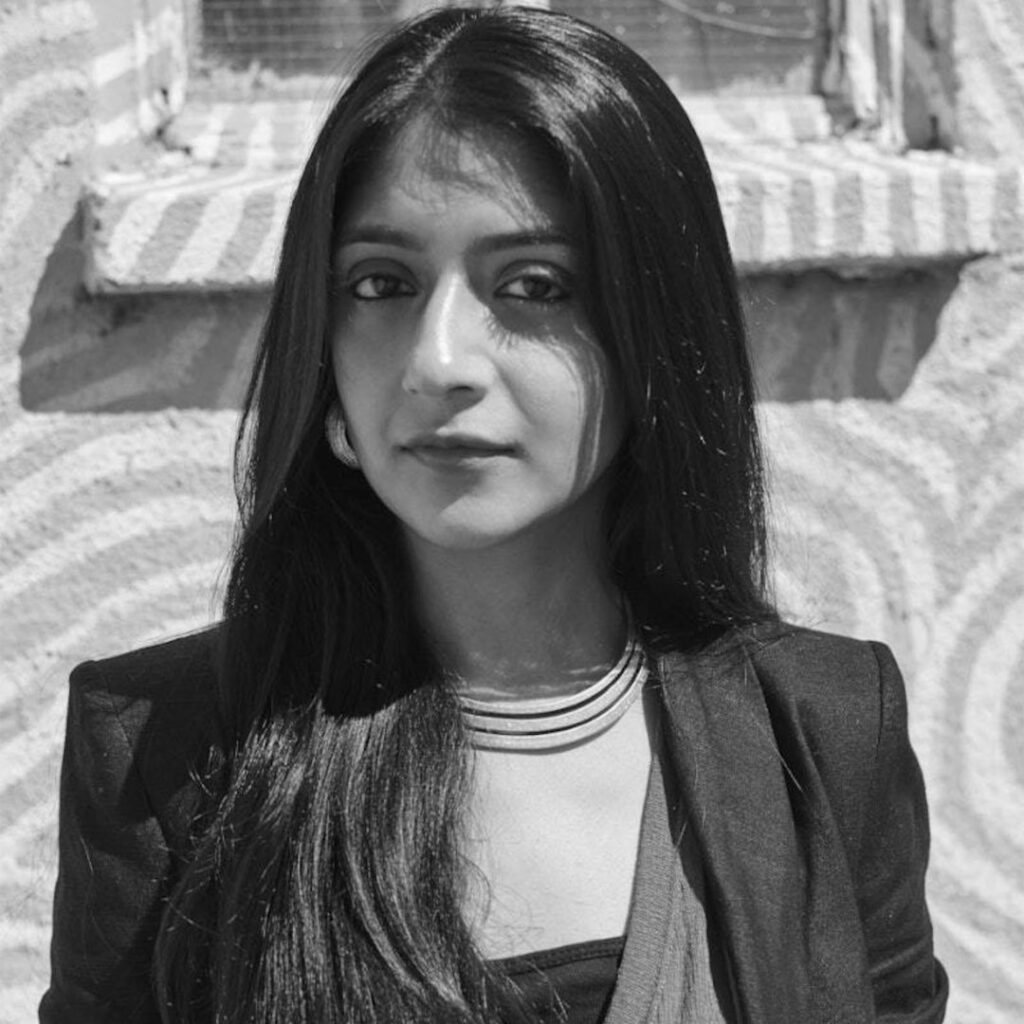 The image is a black and white photograph of Laraib Atta, a Pakistani visual effects (VFX) artist. She is wearing a blazer over a top with horizontal stripes and standing against a background featuring geometric patterns. Laraib Atta is known for her work in Hollywood films such as “10,000 BC,” “The Chronicles of Narnia,” “Prince of Persia,” “Godzilla,” and "X-Men: Days of Future Past"