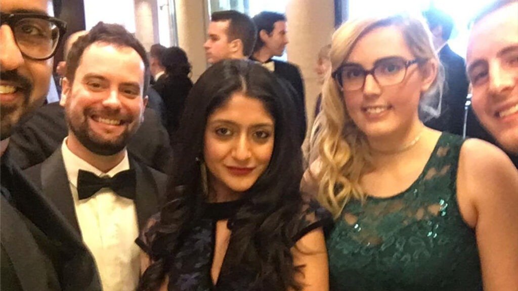 The image shows Laraib Atta, a Pakistani visual effects (VFX) artist, standing in front of a backdrop with a logo and text, likely at a formal event or award ceremony. She is wearing a dark-colored dress and has long, dark hair. Her facial features are clearly visible, and she is smiling at the camera.