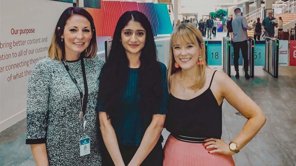 The image shows Laraib Atta, a Pakistani visual effects (VFX) artist, standing with two other individuals in a public area, possibly within a building such as an airport or conference center. The background features a sign with text about bringing better content and connection to customers, and there are turnstile gates with numbers above them.