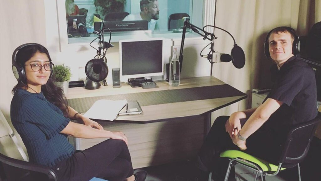 The image shows Laraib Atta, a Pakistani visual effects (VFX) artist, seated opposite an interviewer in a studio setup. Both individuals are in front of microphones, with headphones and other studio equipment visible on the table. The background includes computer monitors and speakers, indicating a professional recording environment.