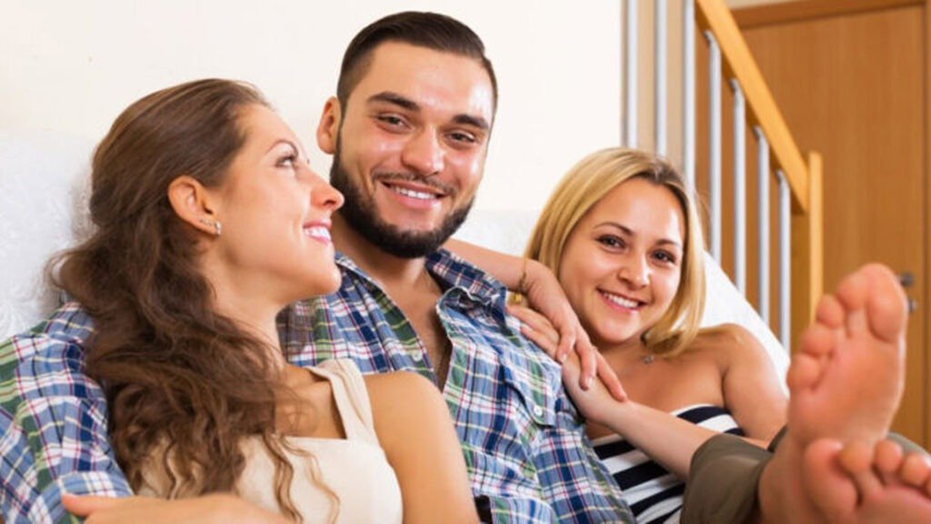 The image depicts three individuals sitting closely together on a staircase represent Symbiosexuality. The person in the center appears to be wearing a plaid shirt, while the individual on the left has long hair and is wearing a dark top. The person on the right is wearing a light-colored top and shorts, with bare feet extended towards the camera.