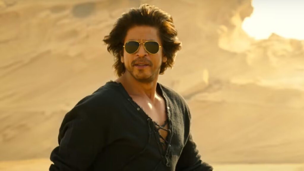 SRK is standing in a desert environment. The person appears to be wearing a dark-colored, long-sleeved shirt with the top few buttons undone. The background consists of a vast expanse of sand and a hazy sky, which suggests a hot and arid climate. This image may be interesting or relevant due to the dramatic setting and the contemplative stance of the figure, which could evoke themes of solitude or adventure.