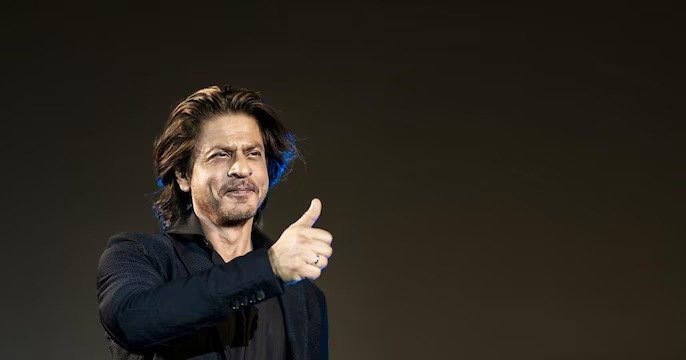 Shah Rukh Khan gives a thumbs-up gesture against a plain, dark background. At Locarno Film Festival