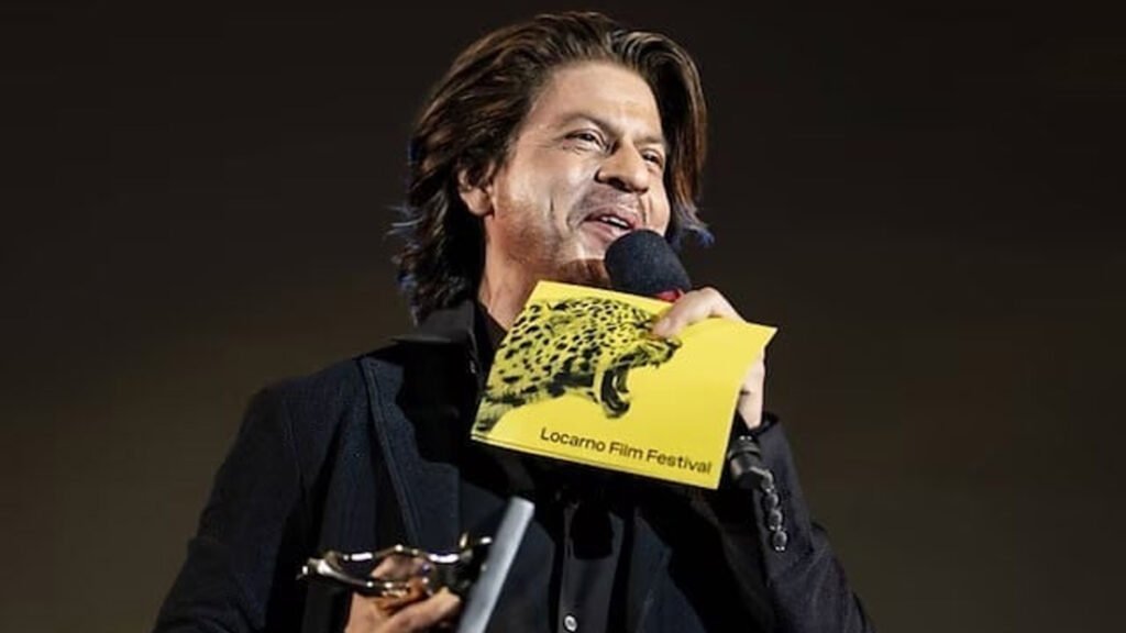 Shah Rukh Khan, stands on a stage holding a microphone and a trophy. They are also holding up a booklet with ‘Locarno Film Festival’ and an image of a leopard on the cover, suggesting the event setting.