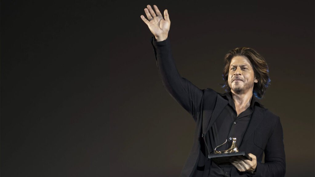 Shah Rukh Khan, dressed in a black suit, stands with his right hand raised in a waving gesture. He holds an award in his left hand. The background features a gradient of dark hues, emphasizing the subject.